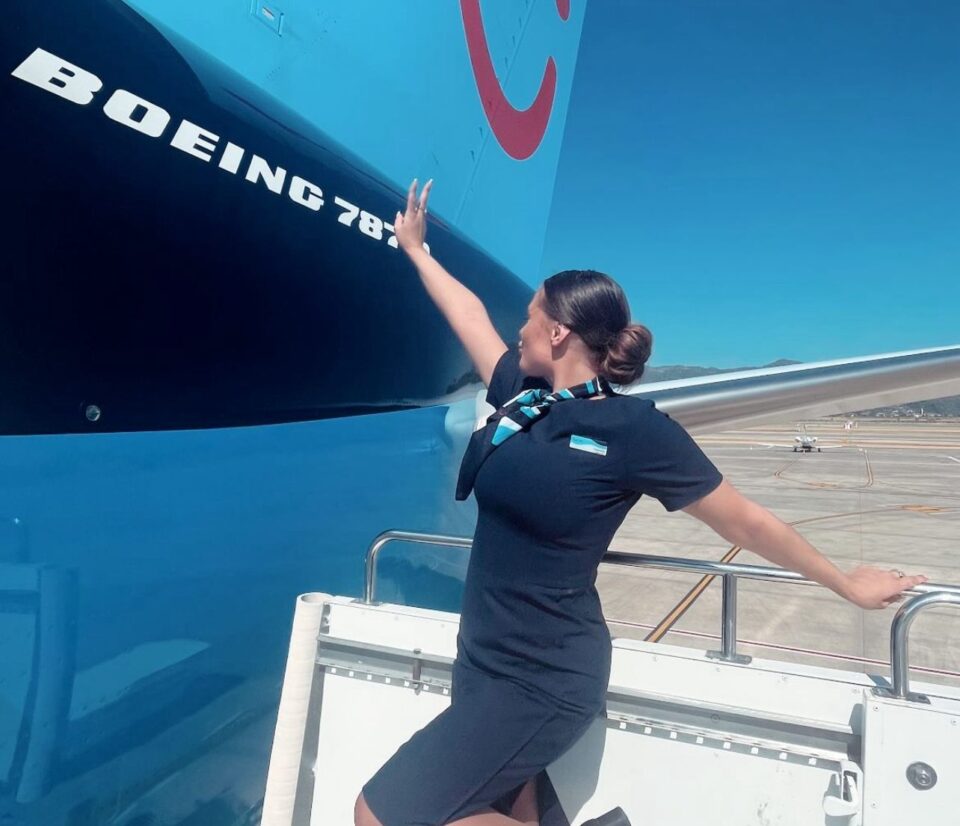 Comissária de bordo posa ao lado de um avião Boeing 787 em pista de aeroporto, vestindo uniforme preto com lenço azul e olhando para a fuselagem da aeronave
