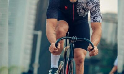 Ciclista homem pedala em alta velocidade por avenida urbana, usando capacete, óculos esportivos e uniforme de ciclismo. Ao fundo, prédios altos e outros ciclistas em movimento.