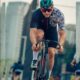 Ciclista homem pedala em alta velocidade por avenida urbana, usando capacete, óculos esportivos e uniforme de ciclismo. Ao fundo, prédios altos e outros ciclistas em movimento.