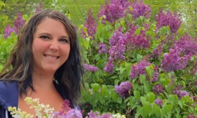Mulher sorri em meio a um jardim florido com arbustos de lilases e flores brancas, em um ambiente ao ar livre cercado por vegetação verde.