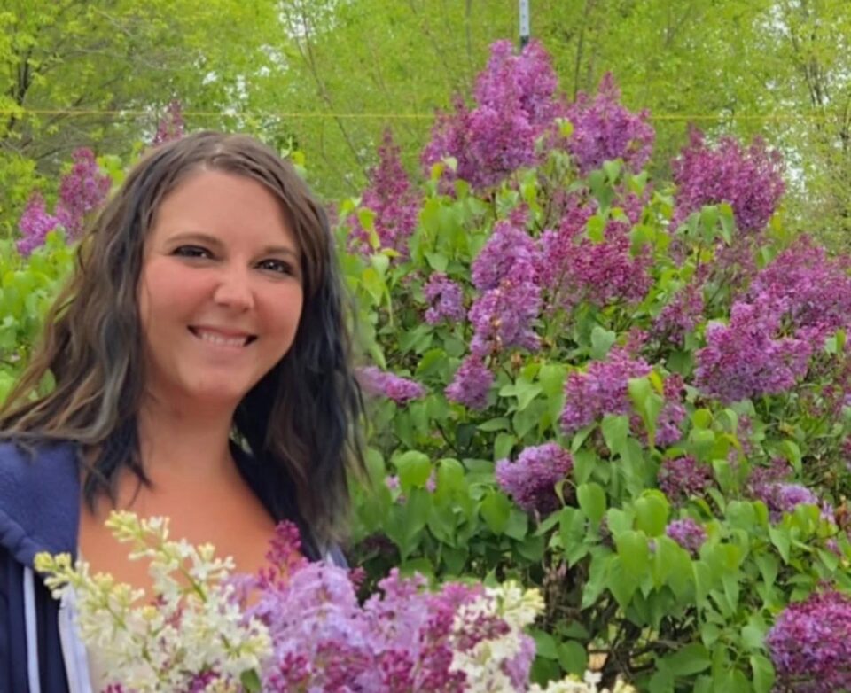 Mulher sorri em meio a um jardim florido com arbustos de lilases e flores brancas, em um ambiente ao ar livre cercado por vegetação verde.