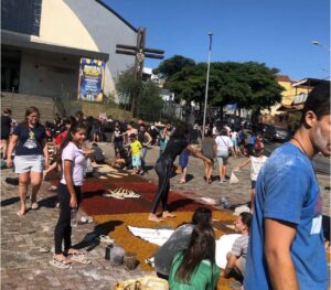 Grupo de pessoas, incluindo crianças e adultos, monta tapetes coloridos de Corpus Christi com serragem e outros materiais em frente a uma igreja, em uma manhã ensolarada. Alguns participantes estão cobertos de pó, e há desenhos religiosos sendo formados no chão
