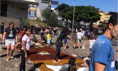 Grupo de pessoas, incluindo crianças e adultos, monta tapetes coloridos de Corpus Christi com serragem e outros materiais em frente a uma igreja, em uma manhã ensolarada. Alguns participantes estão cobertos de pó, e há desenhos religiosos sendo formados no chão