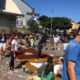 Grupo de pessoas, incluindo crianças e adultos, monta tapetes coloridos de Corpus Christi com serragem e outros materiais em frente a uma igreja, em uma manhã ensolarada. Alguns participantes estão cobertos de pó, e há desenhos religiosos sendo formados no chão