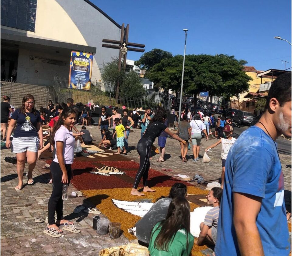 Grupo de pessoas, incluindo crianças e adultos, monta tapetes coloridos de Corpus Christi com serragem e outros materiais em frente a uma igreja, em uma manhã ensolarada. Alguns participantes estão cobertos de pó, e há desenhos religiosos sendo formados no chão
