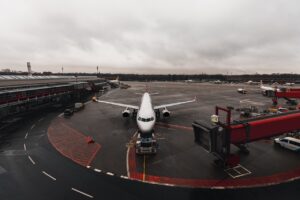 Avião estacionado em frente a um terminal de aeroporto em um dia nublado, com passarela de embarque conectada e veículos de apoio no pátio. A pista está molhada e o céu está coberto por nuvens cinzentas.