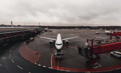 Avião estacionado em frente a um terminal de aeroporto em um dia nublado, com passarela de embarque conectada e veículos de apoio no pátio. A pista está molhada e o céu está coberto por nuvens cinzentas.