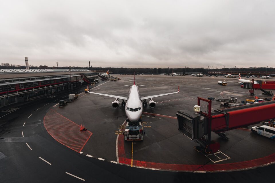 Avião estacionado em frente a um terminal de aeroporto em um dia nublado, com passarela de embarque conectada e veículos de apoio no pátio. A pista está molhada e o céu está coberto por nuvens cinzentas.