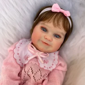 Boneca hiper-realista estilo “bebê reborn” com olhos azuis, cabelo castanho, vestida com roupa rosa de tricô e gola bordada, deitada sobre superfície felpuda branca. Usa faixa com laço rosa na cabeça e tem expressão sorridente.
