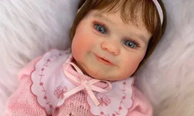 Boneca hiper-realista estilo “bebê reborn” com olhos azuis, cabelo castanho, vestida com roupa rosa de tricô e gola bordada, deitada sobre superfície felpuda branca. Usa faixa com laço rosa na cabeça e tem expressão sorridente.
