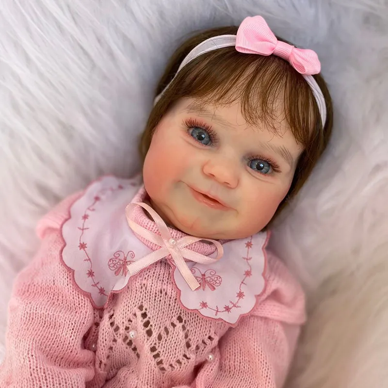 Boneca hiper-realista estilo “bebê reborn” com olhos azuis, cabelo castanho, vestida com roupa rosa de tricô e gola bordada, deitada sobre superfície felpuda branca. Usa faixa com laço rosa na cabeça e tem expressão sorridente.