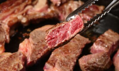 Close em pedaço de carne malpassada sendo segurado por pinça de churrasco; outros cortes aparecem ao fundo, em chapa quente.