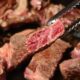 Close em pedaço de carne malpassada sendo segurado por pinça de churrasco; outros cortes aparecem ao fundo, em chapa quente.