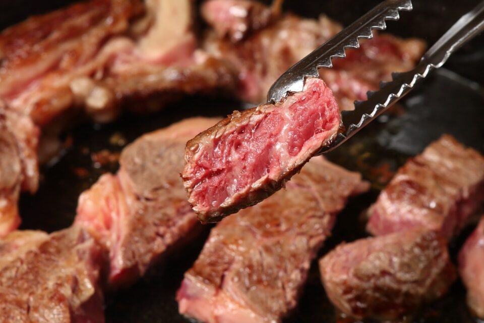 Close em pedaço de carne malpassada sendo segurado por pinça de churrasco; outros cortes aparecem ao fundo, em chapa quente.