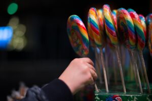 Mão de criança segurando um pirulito grande e colorido entre vários outros, todos dispostos em pé em uma caixa de exposição.