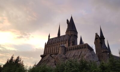 Castelo com arquitetura gótica, semelhante ao de Hogwarts da série Harry Potter, construído sobre uma formação rochosa, cercado por árvores e iluminado por um céu ao entardecer com nuvens coloridas.