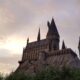 Castelo com arquitetura gótica, semelhante ao de Hogwarts da série Harry Potter, construído sobre uma formação rochosa, cercado por árvores e iluminado por um céu ao entardecer com nuvens coloridas.