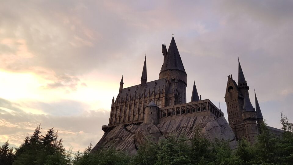 Castelo com arquitetura gótica, semelhante ao de Hogwarts da série Harry Potter, construído sobre uma formação rochosa, cercado por árvores e iluminado por um céu ao entardecer com nuvens coloridas.