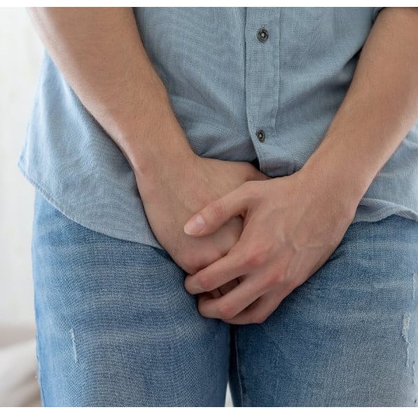 Homem vestido com camisa azul e jeans segura a região genital com as duas mãos, indicando dor ou desconforto