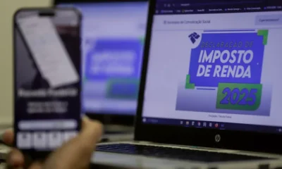 Tela de computador exibe página da Receita Federal com o título "Declaração de Imposto de Renda 2025", enquanto uma mão segura um celular com o aplicativo da Receita aberto em primeiro plano.