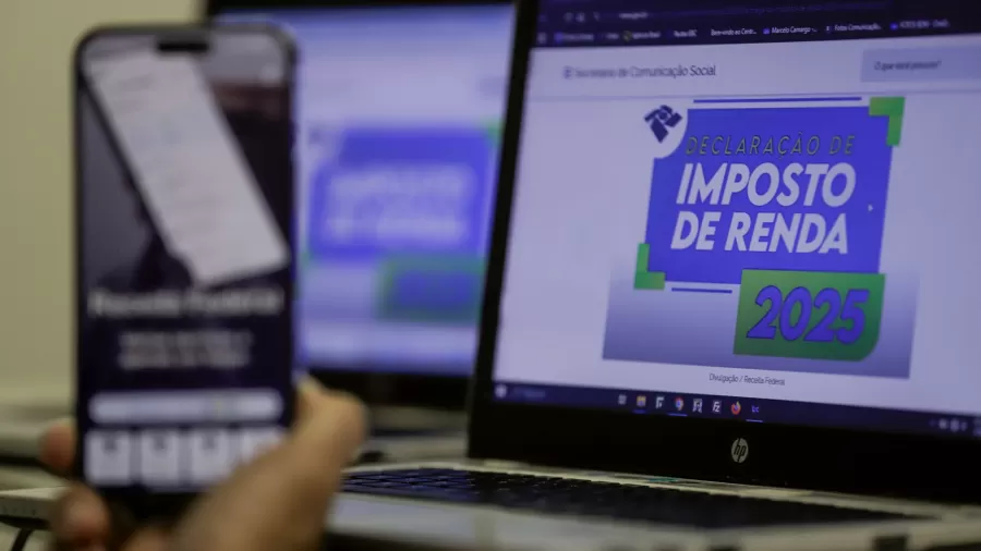 Tela de computador exibe página da Receita Federal com o título "Declaração de Imposto de Renda 2025", enquanto uma mão segura um celular com o aplicativo da Receita aberto em primeiro plano.