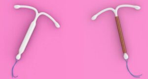 Dois modelos de dispositivo intrauterino (DIU) sobre fundo rosa: à esquerda, um DIU hormonal de plástico branco; à direita, um DIU de cobre com fio espiralado.