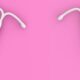 Dois modelos de dispositivo intrauterino (DIU) sobre fundo rosa: à esquerda, um DIU hormonal de plástico branco; à direita, um DIU de cobre com fio espiralado.
