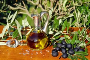 Garrafa de azeite de oliva sobre uma superfície de madeira, cercada por ramos de oliveira e azeitonas pretas, com fundo de vegetação natural