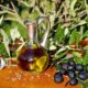 Garrafa de azeite de oliva sobre uma superfície de madeira, cercada por ramos de oliveira e azeitonas pretas, com fundo de vegetação natural