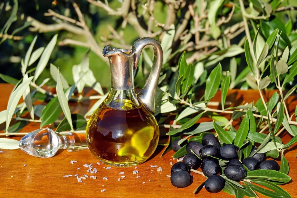 Garrafa de azeite de oliva sobre uma superfície de madeira, cercada por ramos de oliveira e azeitonas pretas, com fundo de vegetação natural