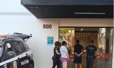 Policiais civis escoltam um homem e uma mulher algemados até a entrada de um prédio da Polícia Civil de São Paulo. Ao lado, viatura preta com o emblema da corporação. Fachada do prédio exibe a sigla DEIC (Departamento Estadual de Investigações Criminais).