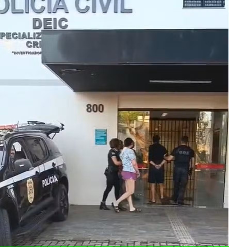 Policiais civis escoltam um homem e uma mulher algemados até a entrada de um prédio da Polícia Civil de São Paulo. Ao lado, viatura preta com o emblema da corporação. Fachada do prédio exibe a sigla DEIC (Departamento Estadual de Investigações Criminais).