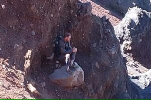 Homem ferido sentado sobre uma pedra, cercado por paredões rochosos, enquanto espera por resgate no Monte Rinjani.