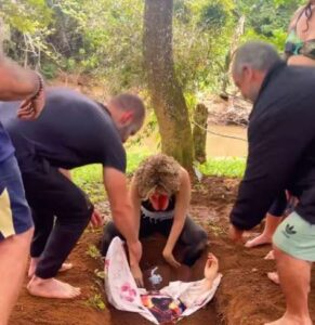 Grupo de pessoas ao redor de uma mulher sendo enterrada até o pescoço em uma cova rasa durante um ritual espiritual ao ar livre, com vegetação ao fundo.