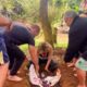 Grupo de pessoas ao redor de uma mulher sendo enterrada até o pescoço em uma cova rasa durante um ritual espiritual ao ar livre, com vegetação ao fundo.