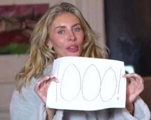 Mulher loira segura cartaz escrito "1000!" com caneta preta; ela veste um roupão cinza e está em um ambiente interno.