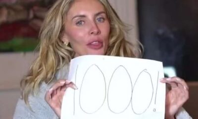 Mulher loira segura cartaz escrito "1000!" com caneta preta; ela veste um roupão cinza e está em um ambiente interno.