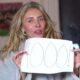 Mulher loira segura cartaz escrito "1000!" com caneta preta; ela veste um roupão cinza e está em um ambiente interno.