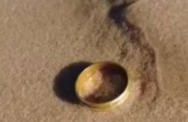 Aliança dourada parcialmente enterrada na areia úmida, com rastro de água escura ao redor.