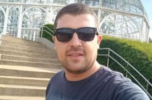 Homem de óculos escuros posando em frente a escadaria de estufa de vidro; identificado como Tiago Nicolau, vítima de ataque fatal em Leme, São Paulo