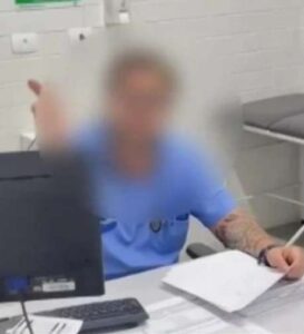 Homem com jaleco azul sentado atrás de uma mesa em consultório médico, com rosto desfocado, segura papéis e gesticula com a mão esquerda.