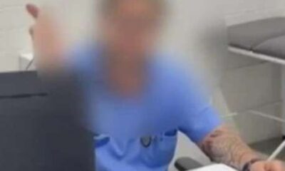 Homem com jaleco azul sentado atrás de uma mesa em consultório médico, com rosto desfocado, segura papéis e gesticula com a mão esquerda.