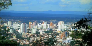 Panorama urbano da cidade de Montes Claros, em Minas Gerais, com prédios altos cercados por vegetação e montanhas ao fundo, sob céu azul com nuvens.