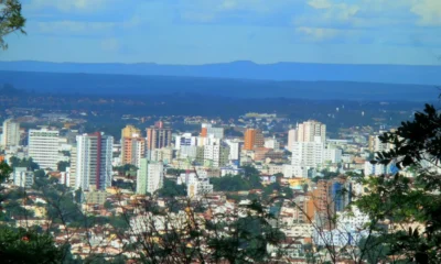 Panorama urbano da cidade de Montes Claros, em Minas Gerais, com prédios altos cercados por vegetação e montanhas ao fundo, sob céu azul com nuvens.