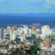 Panorama urbano da cidade de Montes Claros, em Minas Gerais, com prédios altos cercados por vegetação e montanhas ao fundo, sob céu azul com nuvens.