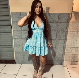 Mulher de cabelos longos e escuros, usando vestido azul claro estampado e sandálias, posa sorrindo em frente a uma parede de azulejos cinza.