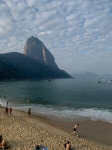 Praia Vermelha no Rio de Janeiro com banhistas na areia e no mar; ao fundo, o Pão de Açúcar sob céu parcialmente coberto por nuvens.