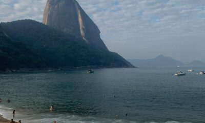 Praia Vermelha no Rio de Janeiro com banhistas na areia e no mar; ao fundo, o Pão de Açúcar sob céu parcialmente coberto por nuvens.