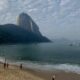 Praia Vermelha no Rio de Janeiro com banhistas na areia e no mar; ao fundo, o Pão de Açúcar sob céu parcialmente coberto por nuvens.