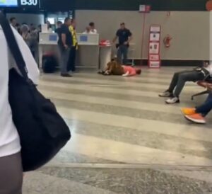 Mulher deitada no chão de um aeroporto, cercada por passageiros e funcionários, em frente ao portão B30 no terminal de embarque
